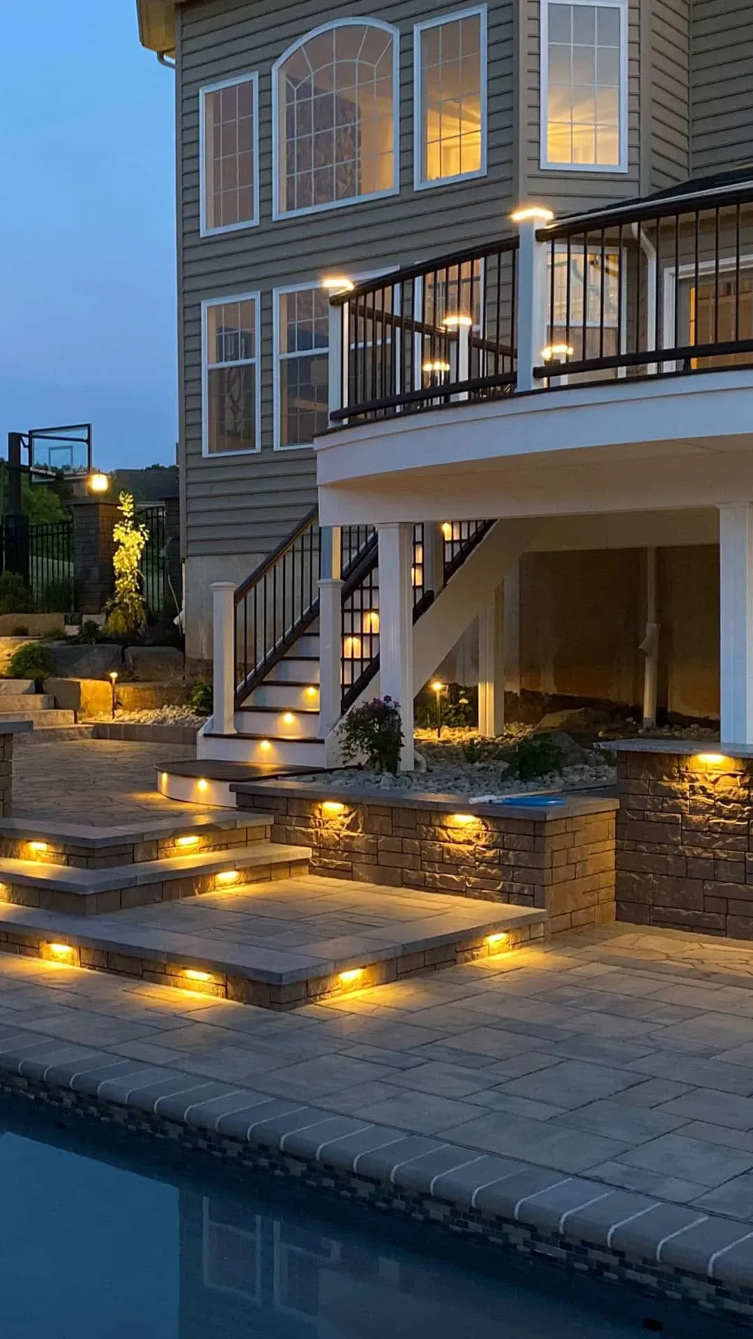 Curved deck with riser and under-cap lighting beside a pool at dusk