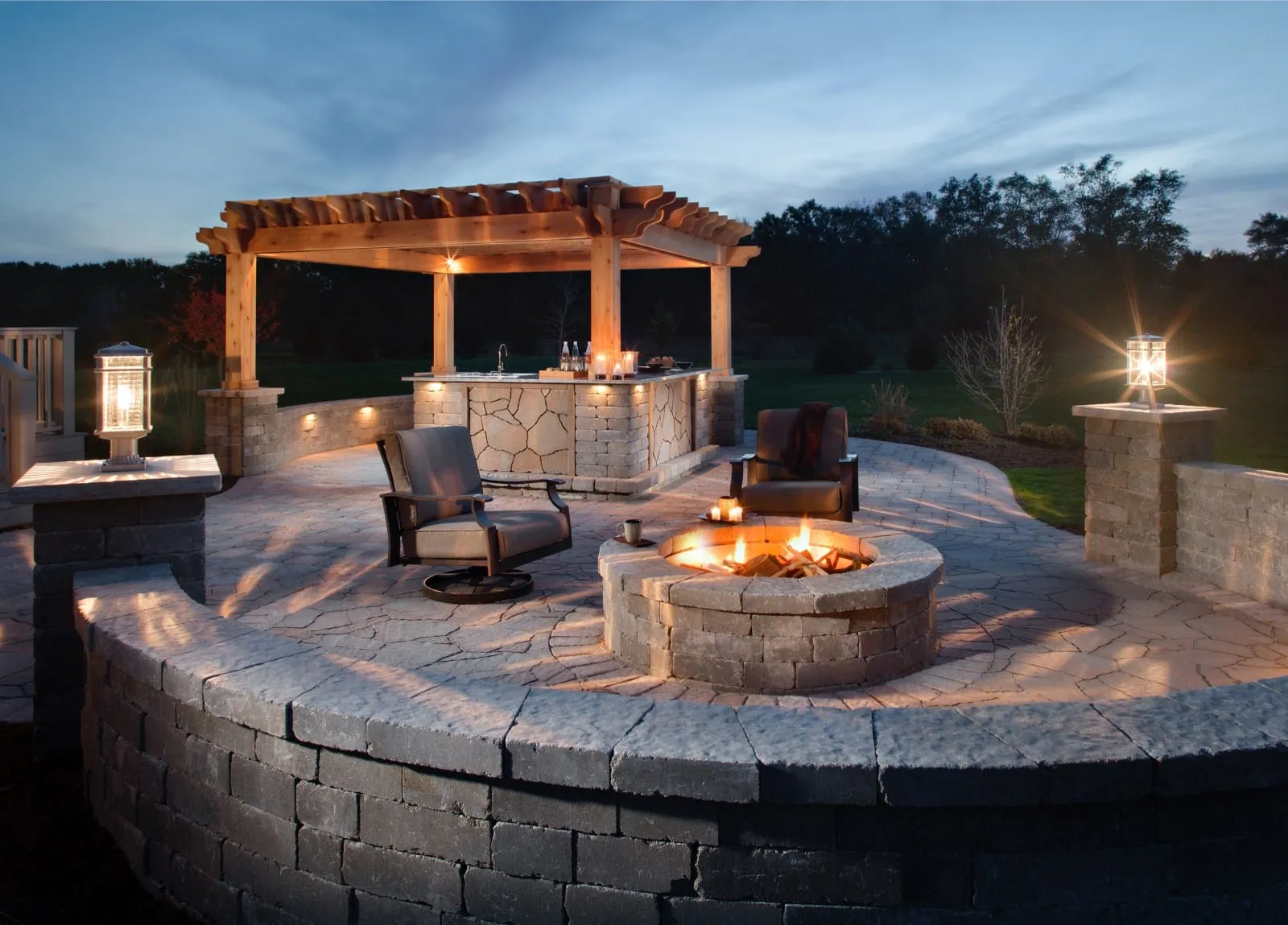 Curved paver patio with pergola, stone outdoor bar, and gas fire pit at dusk